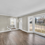 An empty living room in an older home, with bare hardwood floors and a bay window looking onto a quiet residential street