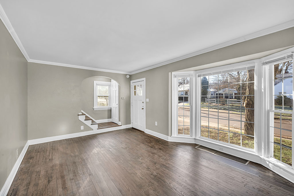An empty living room in an older home, with bare hardwood floors and a bay window looking onto a quiet residential street